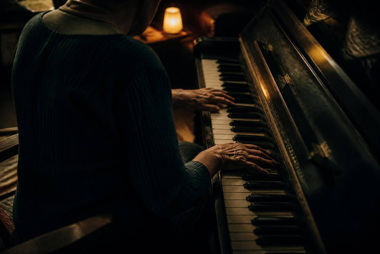 An older person's hands on the keys of an upright piano in a dim room at dusk